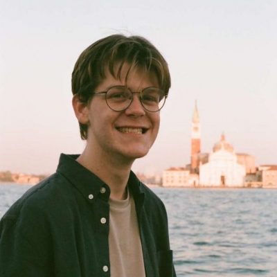 Early 20s white male with blonde hair and wire frame glasses smiling in front of venetian canal and church