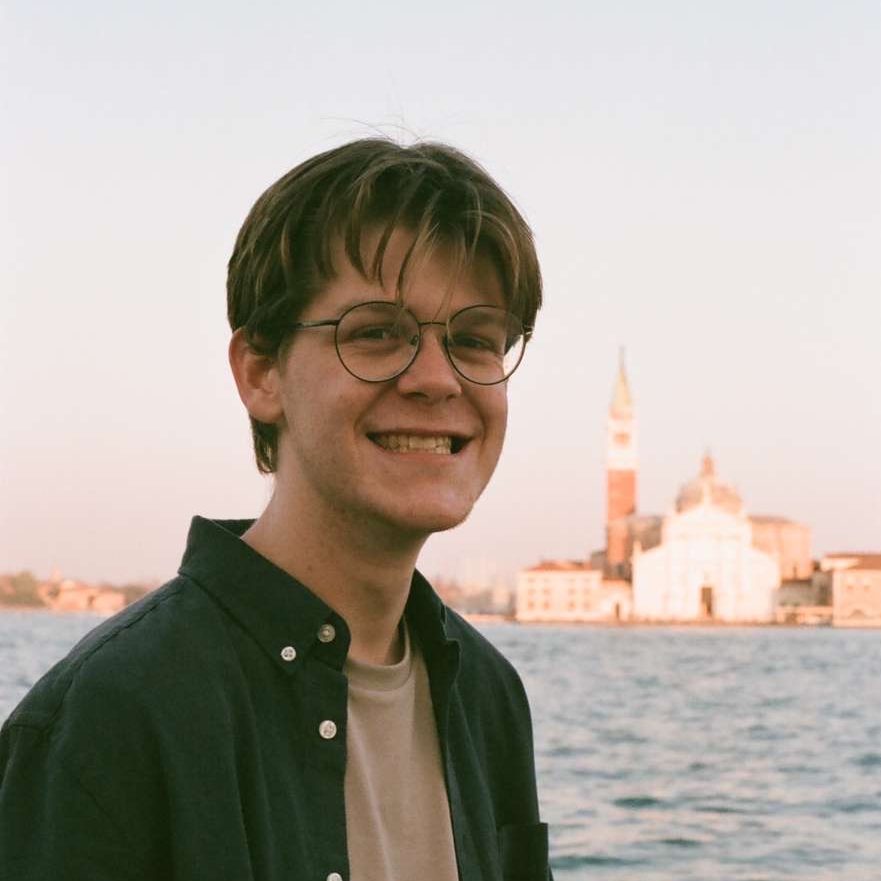 Early 20s white male with blonde hair and wire frame glasses smiling in front of venetian canal and church.