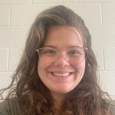 Smiling at camera, middle aged white woman with brown curly hair and rose gold glasses.