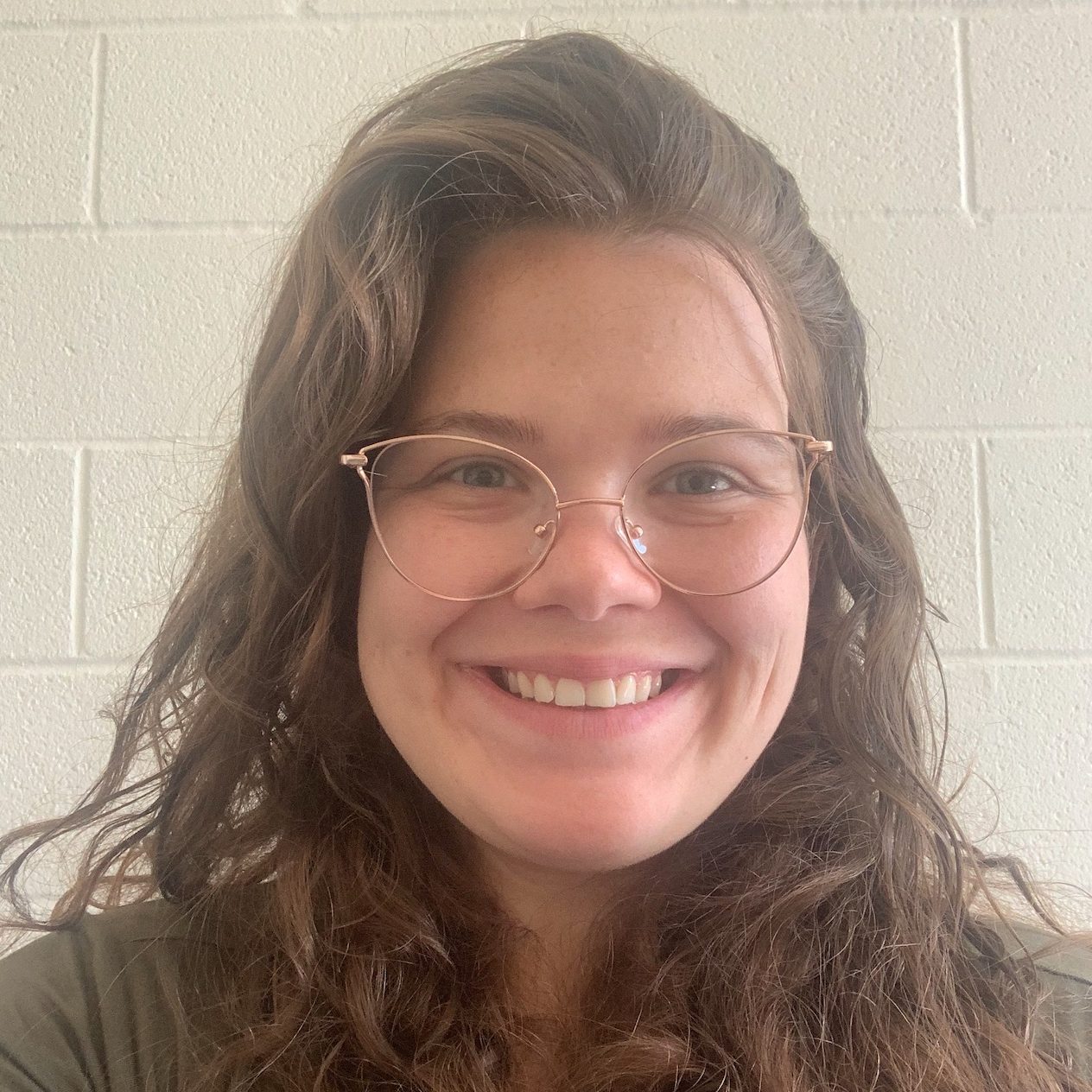 Smiling at camera, middle aged white woman with brown curly hair and rose gold glasses.