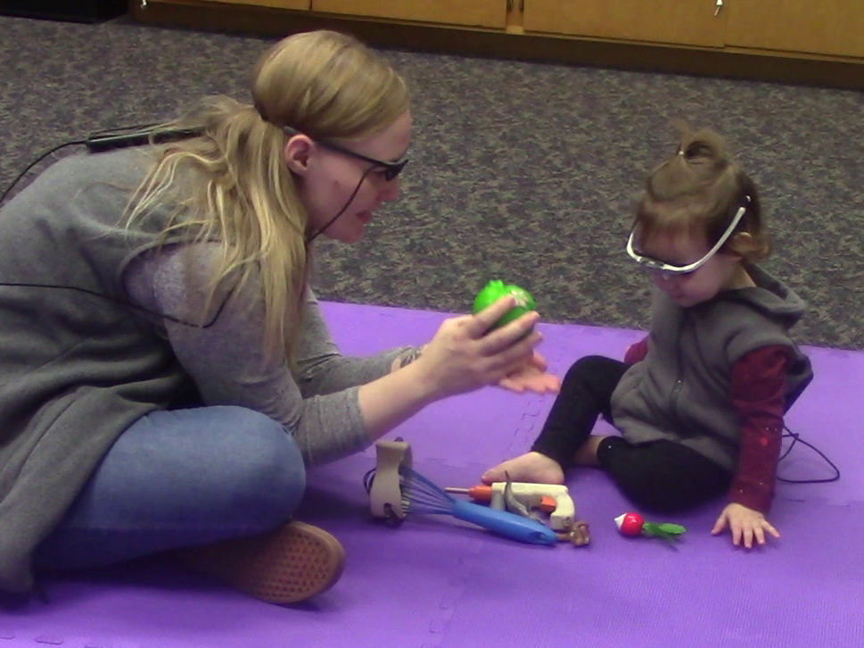 Mother plays with daughter with a variety of toys while wearing eye trackers. Mother holds item up to child's face.