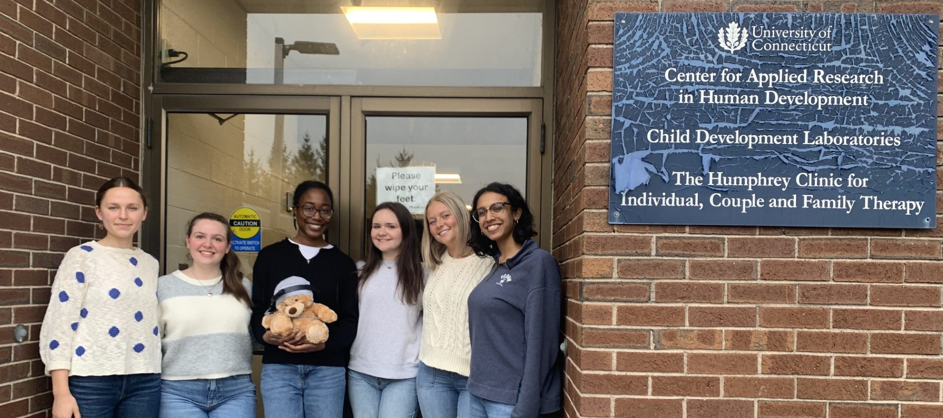 Undergraduate RAs, Juliette, Madeline, Naya, Mackenzie, Adriana, and Sofia (left to right), smile in front of the external door to the Human Development Center.