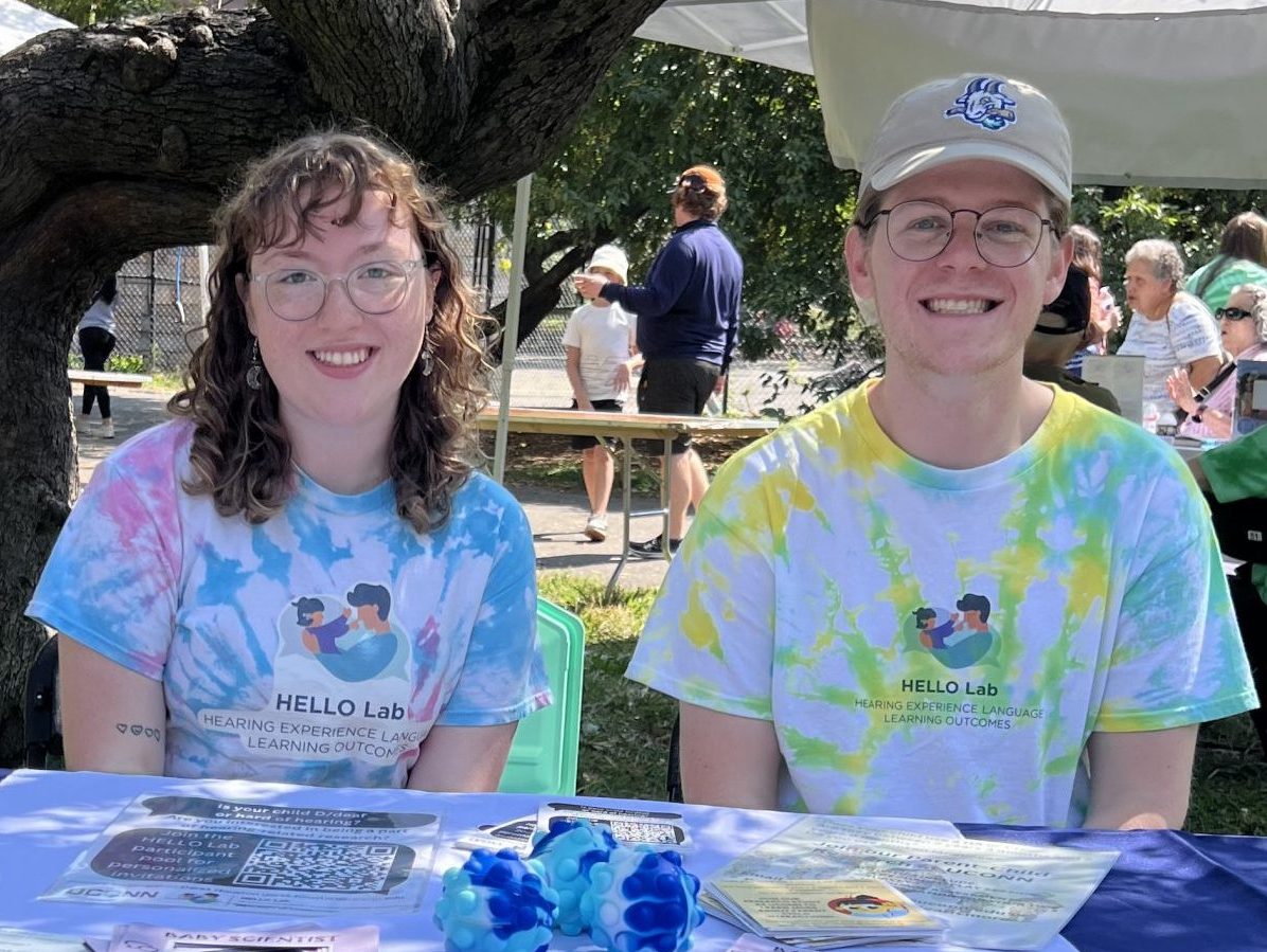 Undergraduate RA, Giovanna, and lab manager, Bobby, smile at a table with a HELLO Lab banner at a recruitment event in NYC.