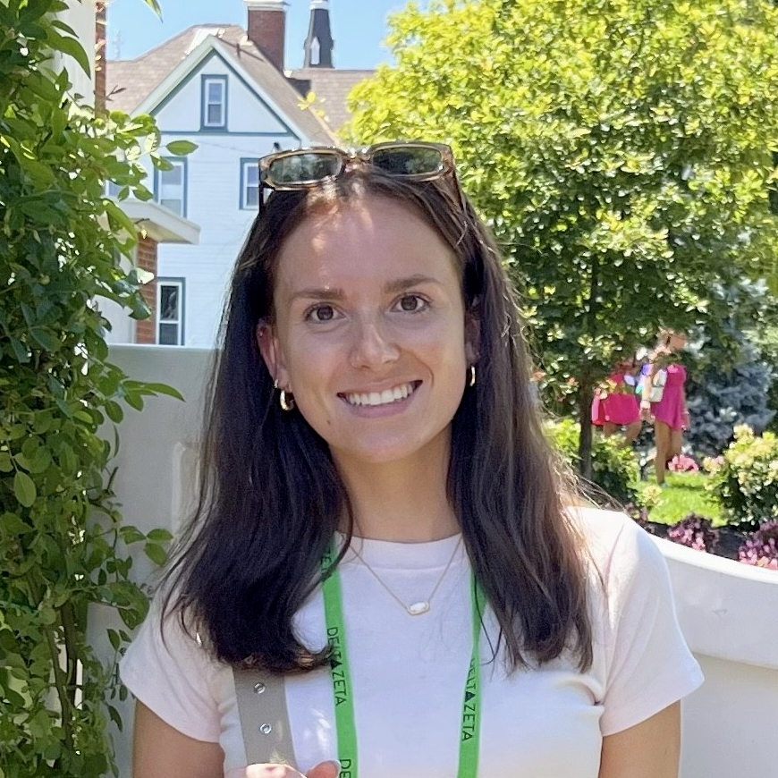 Olivia (a near 20s white woman) smiles wearing a name tag and white t-shirt in front of a decorative gate.